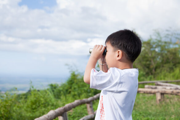 Portrait of little asian boy looking through a telescope, outdoor in the midst of sun light and sky slightly clouded or fair in holiday